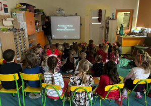 Dzieci siedzą na dywanie i krzesełkach z twarzami skierowanymi w stronę ekranu. Jest to projekcja Mikołajkowego Czytania.